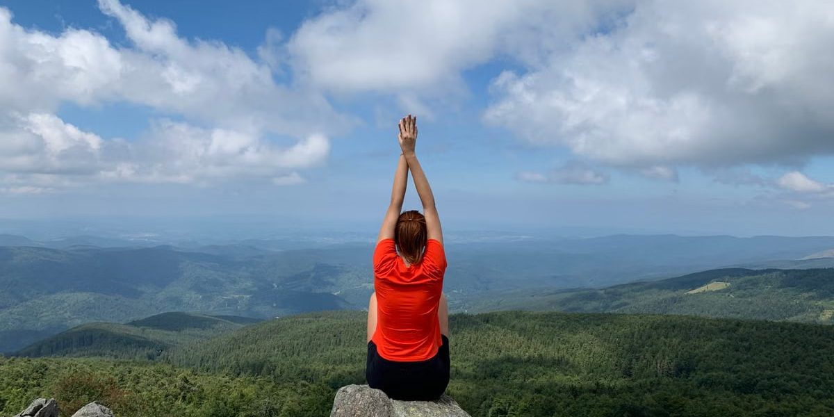 A woman in a red shirt relaxes on a mountain peak with arms raised, enjoying the scenic view.