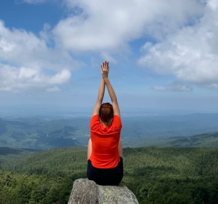 A woman in a red shirt relaxes on a mountain peak with arms raised, enjoying the scenic view.
